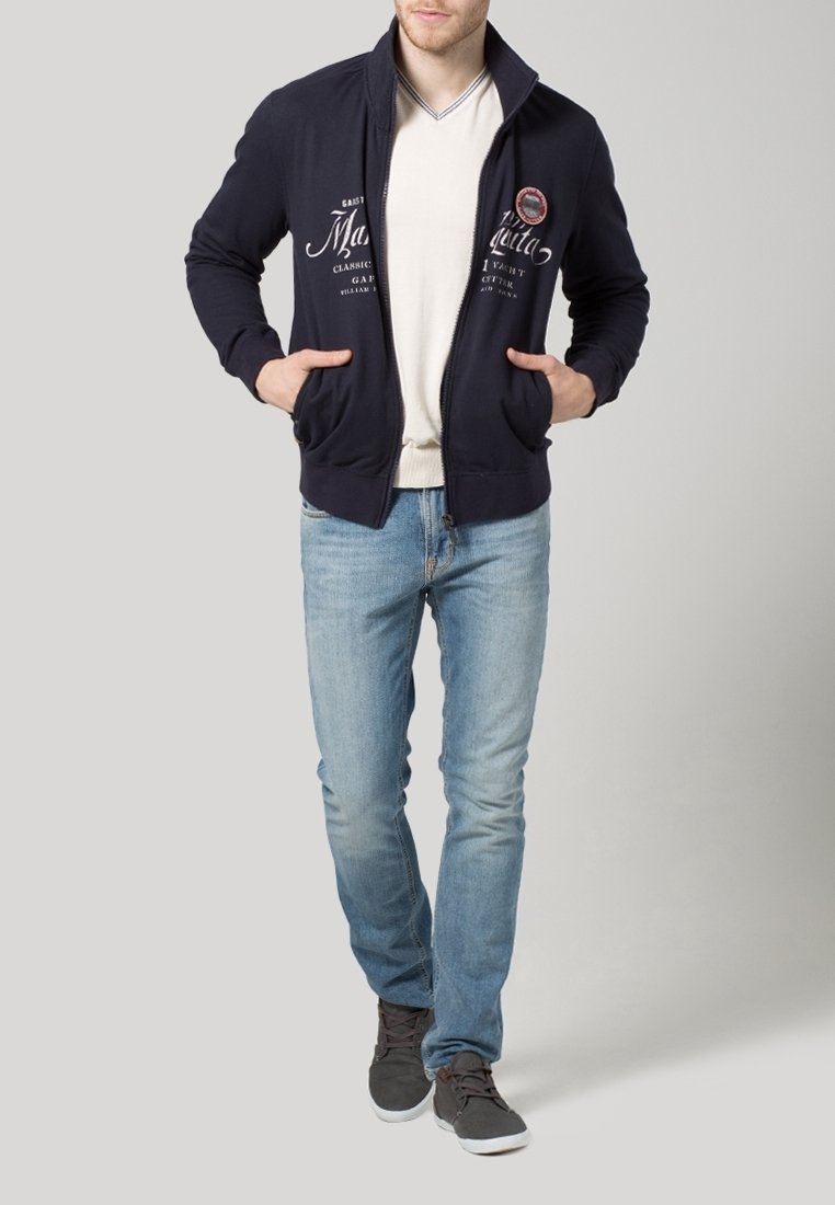 Marinblå zip-up jacka med hög krage, justerbara framfickor och vit grafisk text. Bärs över en ljusbeige skjorta och ljusblå jeans.