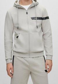 Ljusgrå zip-up hoodie med dragsko på huvan, svart accentrand och framfickor; tillverkad av mjukt material med ribbade ärmslut.