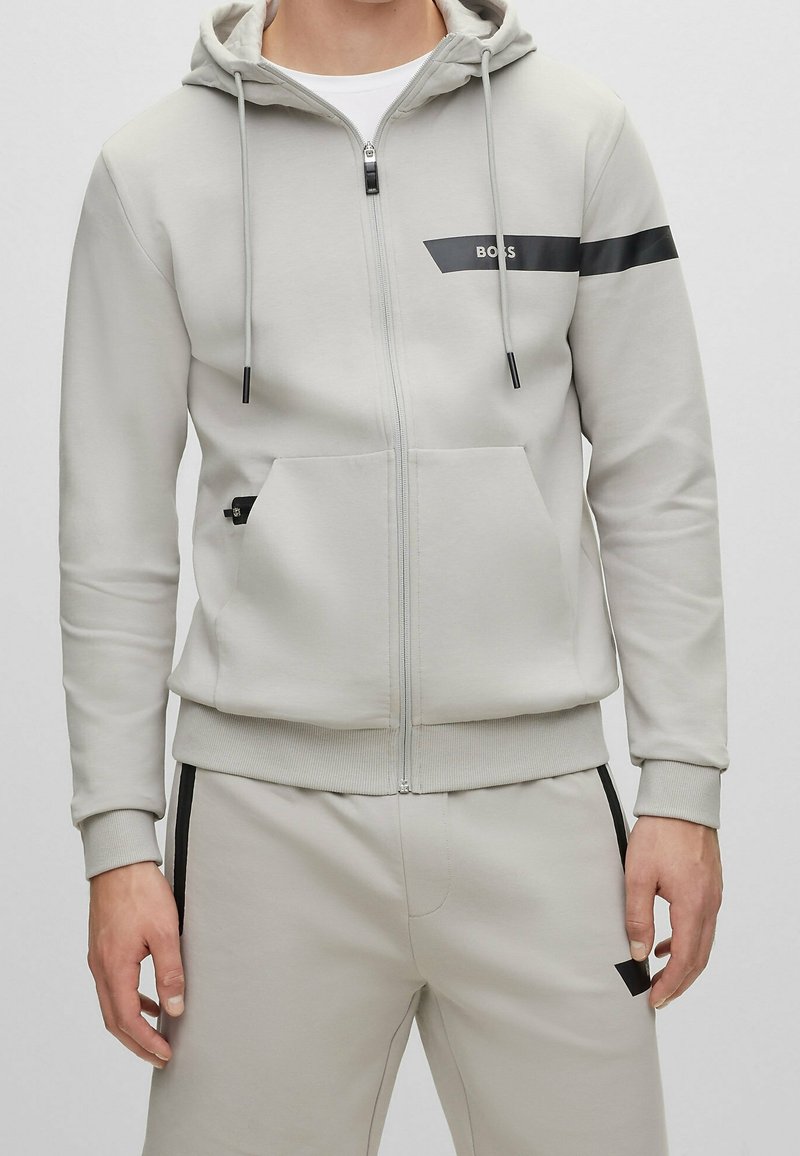 Ljusgrå zip-up hoodie med dragsko på huvan, svart accentrand och framfickor; tillverkad av mjukt material med ribbade ärmslut.