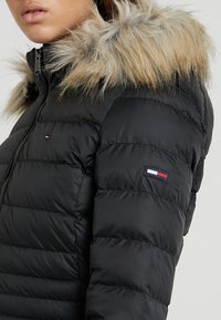 Chaqueta tipo puff negra con capucha de piel sintética. Diseño acolchado horizontal, cierre con cremallera y parche con logo en la manga izquierda. Textura suave.