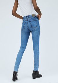 Persona che indossa jeans skinny azzurri chiari e stivaletti neri con tacco, in piedi con le mani sui fianchi su uno sfondo neutro.