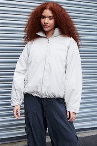 Giacca puffer grigia chiara con colletto alto, zip frontale e polsini elasticizzati. Abbinata a pantaloni cargo scuri. Tessuto strutturato, minimal branding.