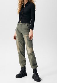 Haut à manches longues texturé noir avec des boutons, associé à un pantalon cargo vert olive avec un patchwork beige et des poches latérales. Bottes à plateforme noires.