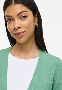 Cardigã verde claro feito de um tecido macio e texturizado; design de frente aberta sobre uma camisa branca. Destaques incluem um decote em V.