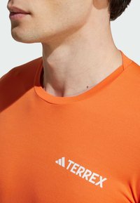 Oranje T-shirt met korte mouwen, gemaakt van een gladde stof, met een ronde hals en een wit "TERREX" logo aan de linkerkant.