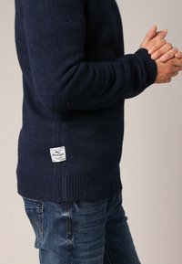 Pull en tricot bleu marine avec un ourlet et des poignets côtelés, portant une petite étiquette blanche sur le côté inférieur gauche. Associé à un jean bleu.