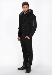 Sort polstret jakke med hætte, lynlåslommer og en glat tekstur. Bæres med sorte bukser og sorte snørebodstøvler. Stående pose.