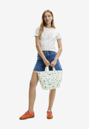 Borsa a rete bianca con pois blu, dotata di due manici. La modella indossa una maglietta bianca, pantaloni corti in denim e sneakers arancioni.