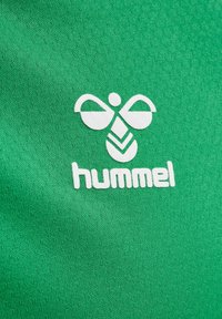 Wit gestileerd bijenlogo en "hummel" tekst op een getextureerde groene stoffen achtergrond, waarschijnlijk sportkledingbranding.