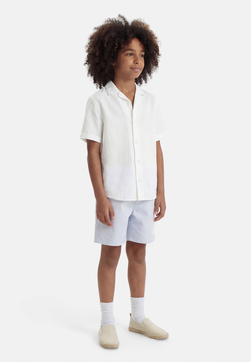 Αγόρι με σγουρά μαλλιά που φοράει λευκό κοντομάνικο πουκάμισο με κουμπιά, ανοιχτόχρωμο μπλε σορτς, λευκές κάλτσες και μπεζ παπούτσια τύπου slip-on, στέκεται ήρεμα.