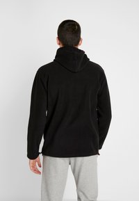 Felpa nera in pile con vestibilità rilassata, cappuccio regolabile con cordino e maniche lunghe. Il materiale ha una consistenza morbida. Indossata con pantaloni della tuta grigi.