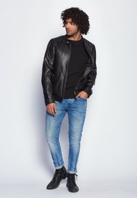 Sort læderjakke med teksturerede paneler, båret over en sort skjorte. Parret med blå slim-fit jeans og sorte snørebåndsstøvler.