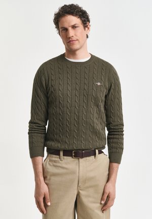 Mann trägt einen olivgrünen Pullover mit Zopfmuster und beigefarbene Hose mit braunem Gürtel, stehend vor einem schlichten hellen Hintergrund.