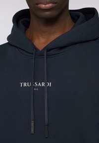 Marinblå hoodie med texturerad tyg. Har dragsnören och en framsida med en logotyp som säger "TRUSSARDI 1911" i vitt. Minimalistisk design.