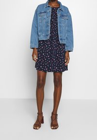 Kurtka dżinsowa na granatowej sukience w kwiaty z małymi białymi i różowymi kwiatami. Noszona z bordowymi sandałami na obcasie z paskami, prezentując swobodny styl.