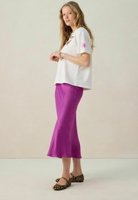 T-shirt blanc à manches courtes avec impression graphique, associé à une jupe satin violette longueur genoux et des chaussures plates à imprimé léopard. Textures lisses et couleurs vives.