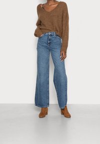 Maglione lavorato a maglia marrone con scollo a V, abbinato a jeans azzurri a vita alta e a gamba larga e stivaletti marroni. Texture liscia e vestibilità casual.