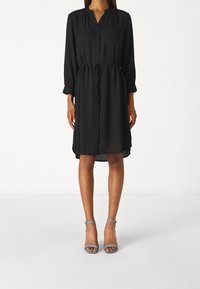 Svart klänning i knälängd med en knäppt krage, långa ärmar, dragsko i midjan och flödande textur. Matchas med ljusblå klacksandaler.