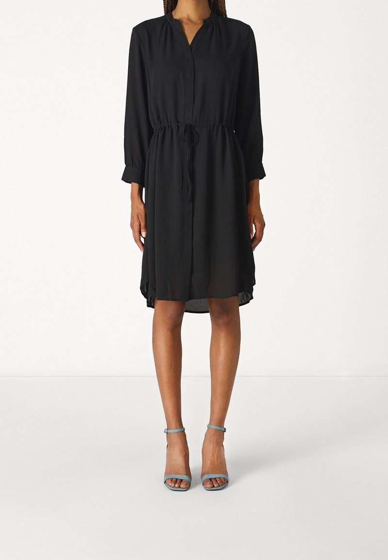 Svart klänning i knälängd med en knäppt krage, långa ärmar, dragsko i midjan och flödande textur. Matchas med ljusblå klacksandaler.