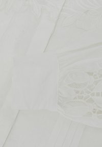 Material alb cu broderie florală și detalii de dantelă decupată, prezentând o prim-plan a manșetei mânecii și modele texturate.