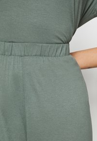 Losvallend jumpsuit met elastische tailleband in een gedempte groene stof, met een zachte textuur en subtiele glans. Zichtbaar detail van een zijzak.