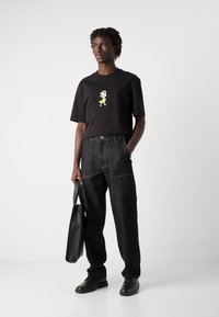 Camiseta negra con un gráfico de dibujos animados en verde, combinada con pantalones vaqueros oscuros holgados. El modelo lleva un bolso tote negro y zapatos negros.