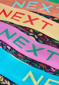 Farebné látkové pásy s výšivkou slova "NEXT" v rôznych farbách na textílii s kvetinovým vzorom.