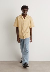 Short-sleeve, light yellow shirt with a textured pattern and black buttons, paired with light blue jeans and black shoes.
