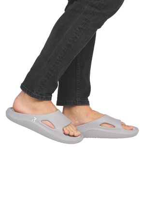 Des sandales grises à enfiler en matériau semblable au caoutchouc, présentant des découpes et une semelle texturée pour l'adhérence, portées avec un jean sombre.