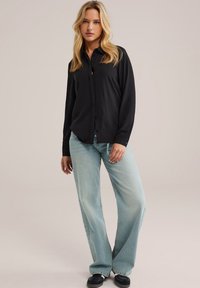 Donna con lunghi capelli biondi, indossa una camicia nera con bottoni, jeans larghi blu chiaro e sneaker nere, in piedi contro uno sfondo neutro.