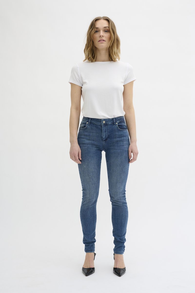 Bela kratka majica s kratkimi rokavi, v kombinaciji s tesnimi temno modrimi skinny jeans hlačami z visokim pasom in minimalnim detajliranjem, obuta v črne špice.