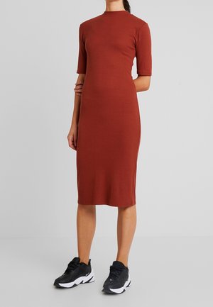 Frau trägt ein figurbetontes, knielanges, rostfarbenes Kleid mit ellenbogenlangen Ärmeln, kombiniert mit schwarzen und weißen Sportschuhen.
