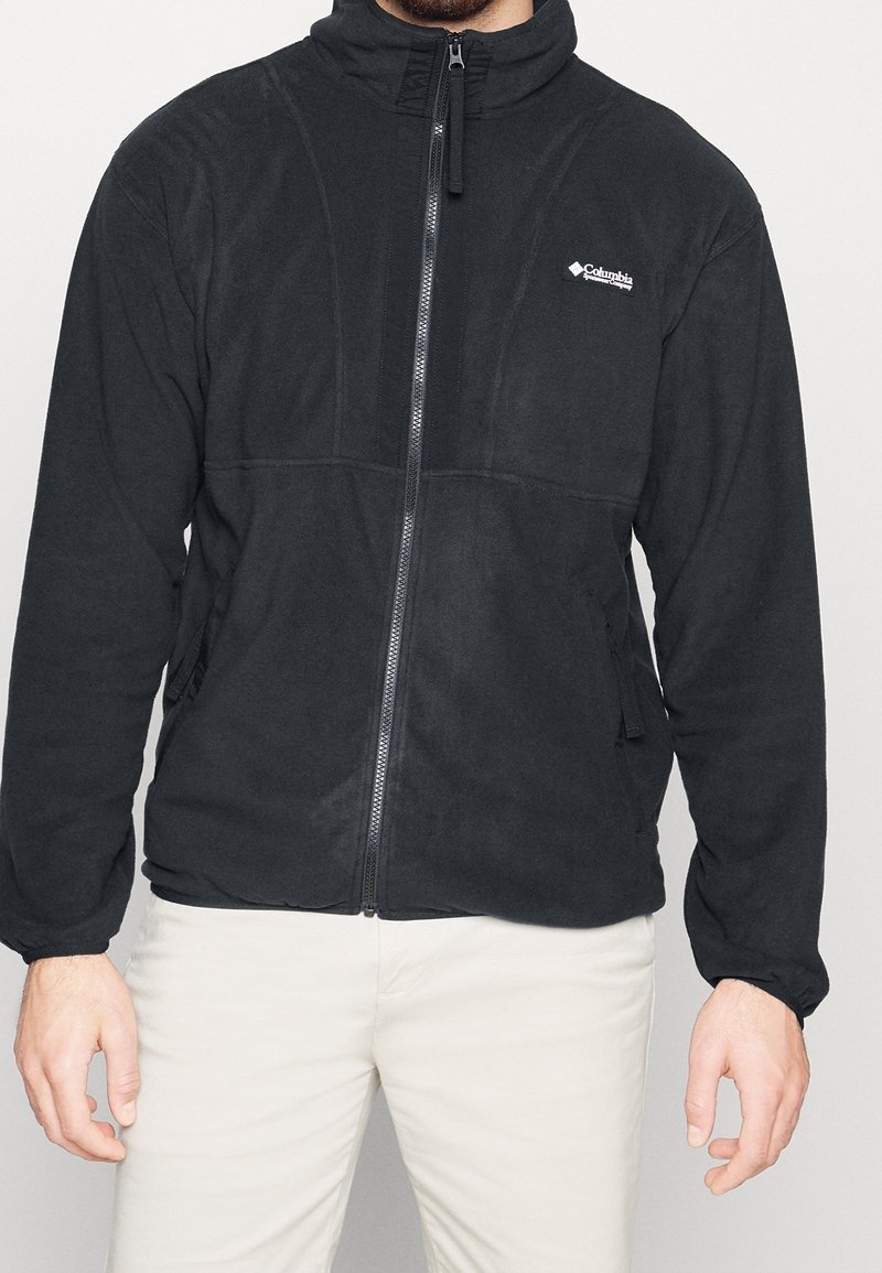 Homme portant une veste polaire noire Columbia zippée et un pantalon de couleur claire, debout devant un fond clair uni.