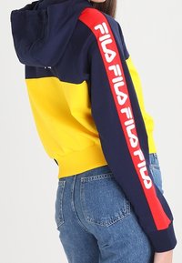 Felpa con cappuccio con design a blocchi di colore in giallo e blu navy, con una striscia rossa sulla manica e logo in caratteri bianchi audaci.
