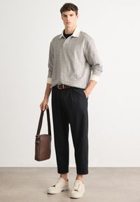 Young man wearing patterned long-sleeve polo, black cropped pants, beige sneakers, and holding a brown shoulder bag against a plain background.