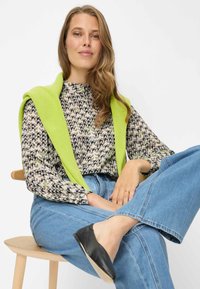 Blusa con motivo ondeggiato nero e beige, abbinata a jeans blu e maglione verde lavorato a maglia drappeggiato sulle spalle. Scarpe basse nere.