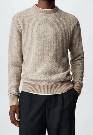 Homme portant un pull en tricot beige texturé sur une chemise blanche, associé à un pantalon noir, se tenant avec une main dans la poche.