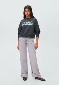 Giovane donna con lunghi capelli scuri, che indossa una felpa scura con il testo "TEAM BLONDE", jeans grigio chiaro e scarpe nere con borchie, in piedi.