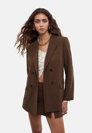 Blazer marron à double boutonnage en tissu lisse, avec des boutons noirs et des revers crantés, porté sur un haut en dentelle clair et un short assorti.