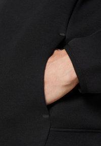 Svart sweatshirt-tyg med en ficka och en hand vilande inuti. Texturen är slät, med en enkel och diskret design.