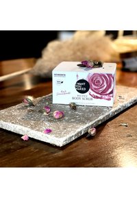 Scatola di scrub per il corpo con design a rose, cartone bianco, etichetta vegana. Posizionata su una superficie di pietra texturizzata con petali di rosa secchi sparsi.