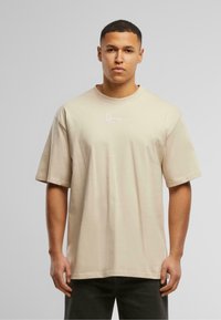 Khaki oversized t-shirt with short sleeves, round neckline, and white embroidered logo on the chest, made from soft, lightweight fabric.