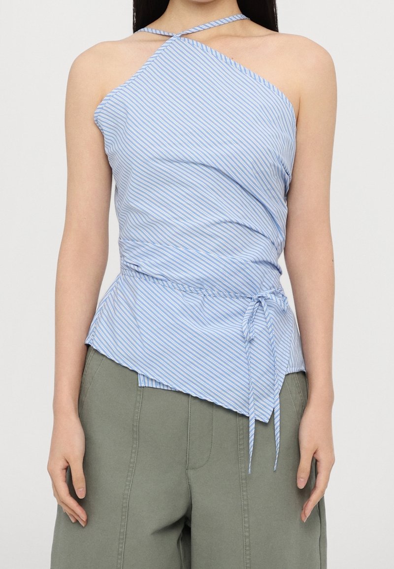 Femme portant un top à bretelles rayé bleu et blanc avec un ourlet asymétrique et une taille nouée, associé à un pantalon ample vert olive.