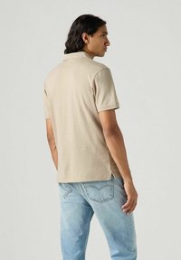 Beige pikétröja med korta ärmar, sidsprickor vid fållen, bärs med ljusblå jeans, vilket visar en avslappnad passform och mjuk textur.