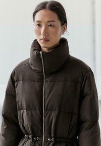 Brązowa, pikowana kurtka z wysokim kołnierzem i elastycznym ściągaczem w talii. Posiada przedni zamek błyskawiczny i gładkie, matowe wykończenie.