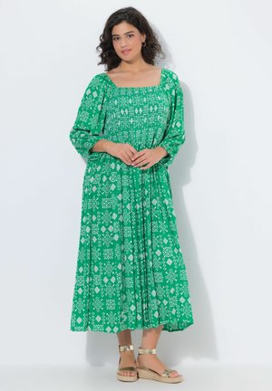 Kobieta ubrana w zieloną, wzorzystą sukienkę midi z długim rękawem, kwadratowym dekoltem i złotymi sandałami na koturnie, stojąca na białym tle.