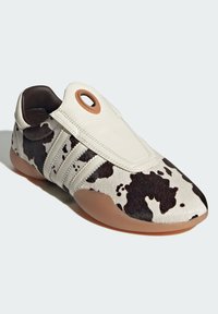 Sneaker featuring a white leather upper with black cow print, tan rubber sole, and a distinctive circular accent at the heel.