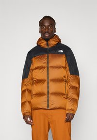 Jachetă puffer în nuanță de portocaliu ars, cu accente negre, având închidere cu fermoar, glugă și design matlasat. Material din nailon cu manșete elastice.