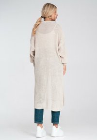 Długi, jasnobeżowy kardigan z dzianiny o luźnym kroju, z opadającymi ramionami, ribbowymi mankietami i strukturalnym wzorem, noszony na niebieskich dżinsach i białych trampkach.