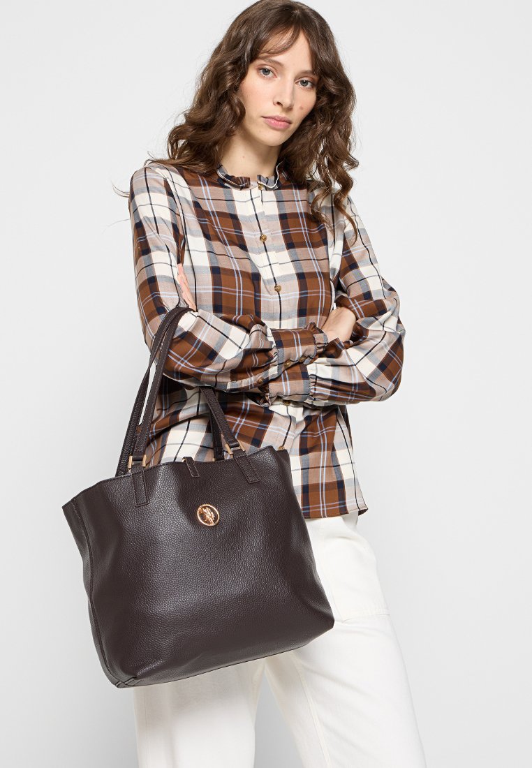 Braune Ledertasche mit strukturiertem Finish, zwei stabilen Griffen und einem runden Logo-Akzent. Trägt man zusammen mit einem karierten Hemd.